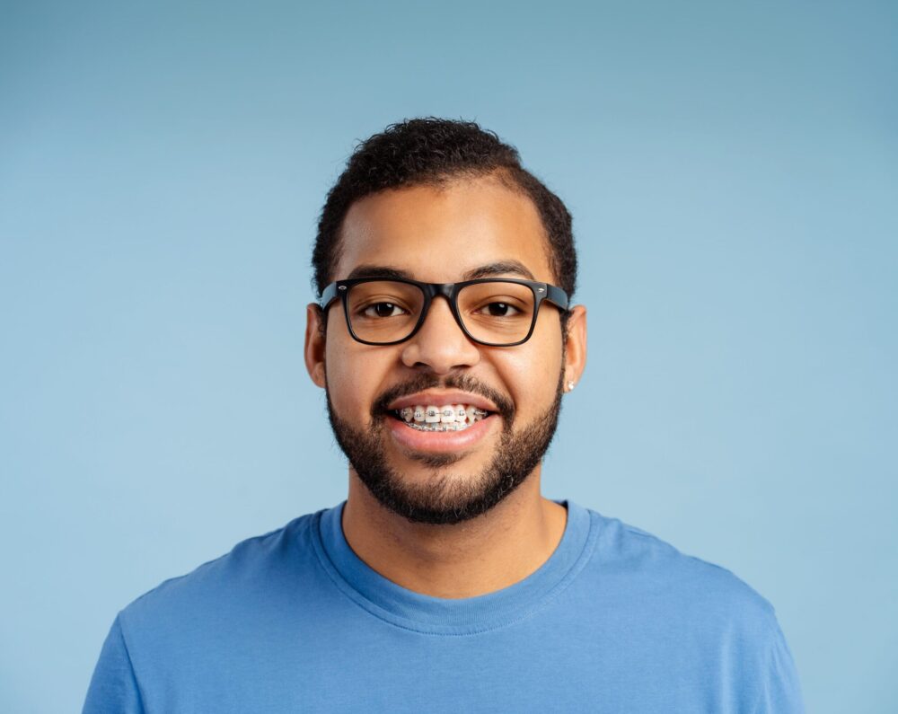 Smiling male with braces in a casual t-shirt and glasses Smiling man wearing braces, proudly showing the journey to a healthier, more confident smile through the benefits of modern orthodontic treatment.
