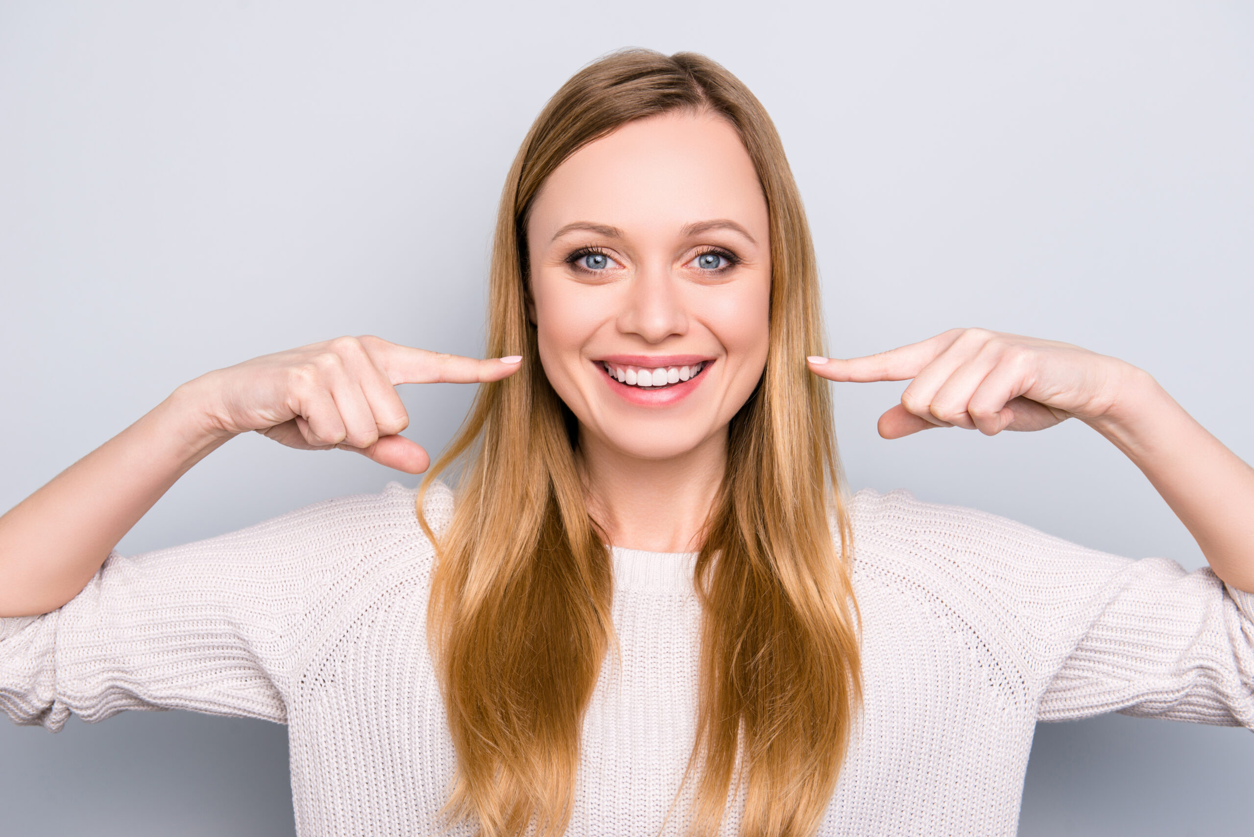 Portrait of joyful satisfied girl gesturing her beaming white healthy teeth with two forefingers looking at camera isolated on grey background. Orthodontic concept Smiling woman pointing at her teeth with both hands, proudly showing off the beautiful, healthy results achieved through orthodontic treatment.