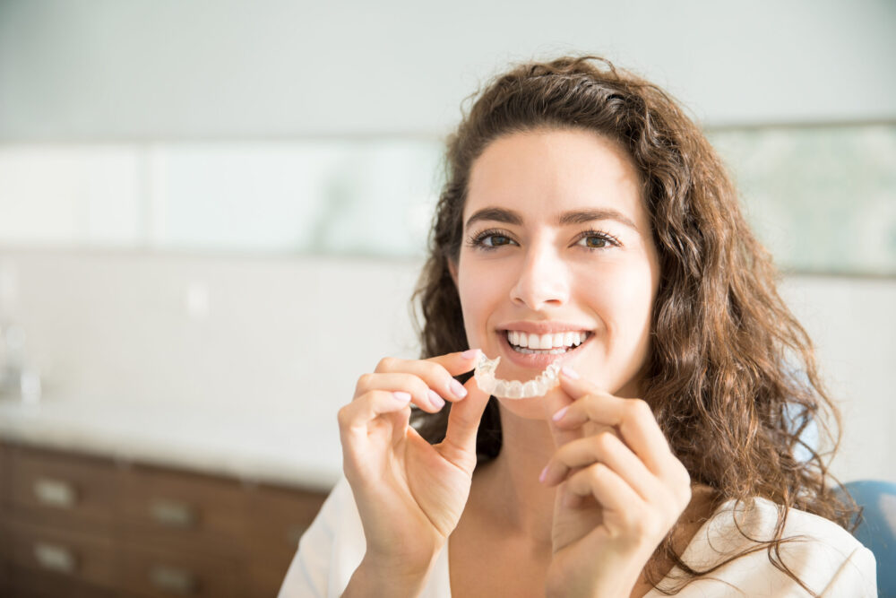 Adult Patient Holding Orthodontic Aligners Woman smiling as she removes clear aligners, highlighting the discreet and attractive way modern orthodontics enhances her smile’s appearance.